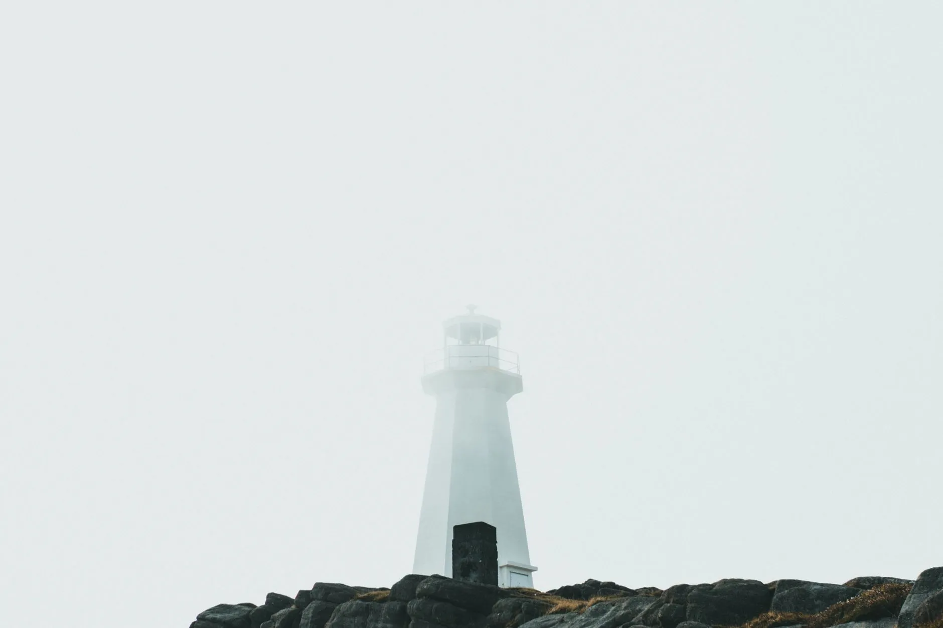 Menara mercusuar muncul samar dari kabut di tepi pantai berbatu.