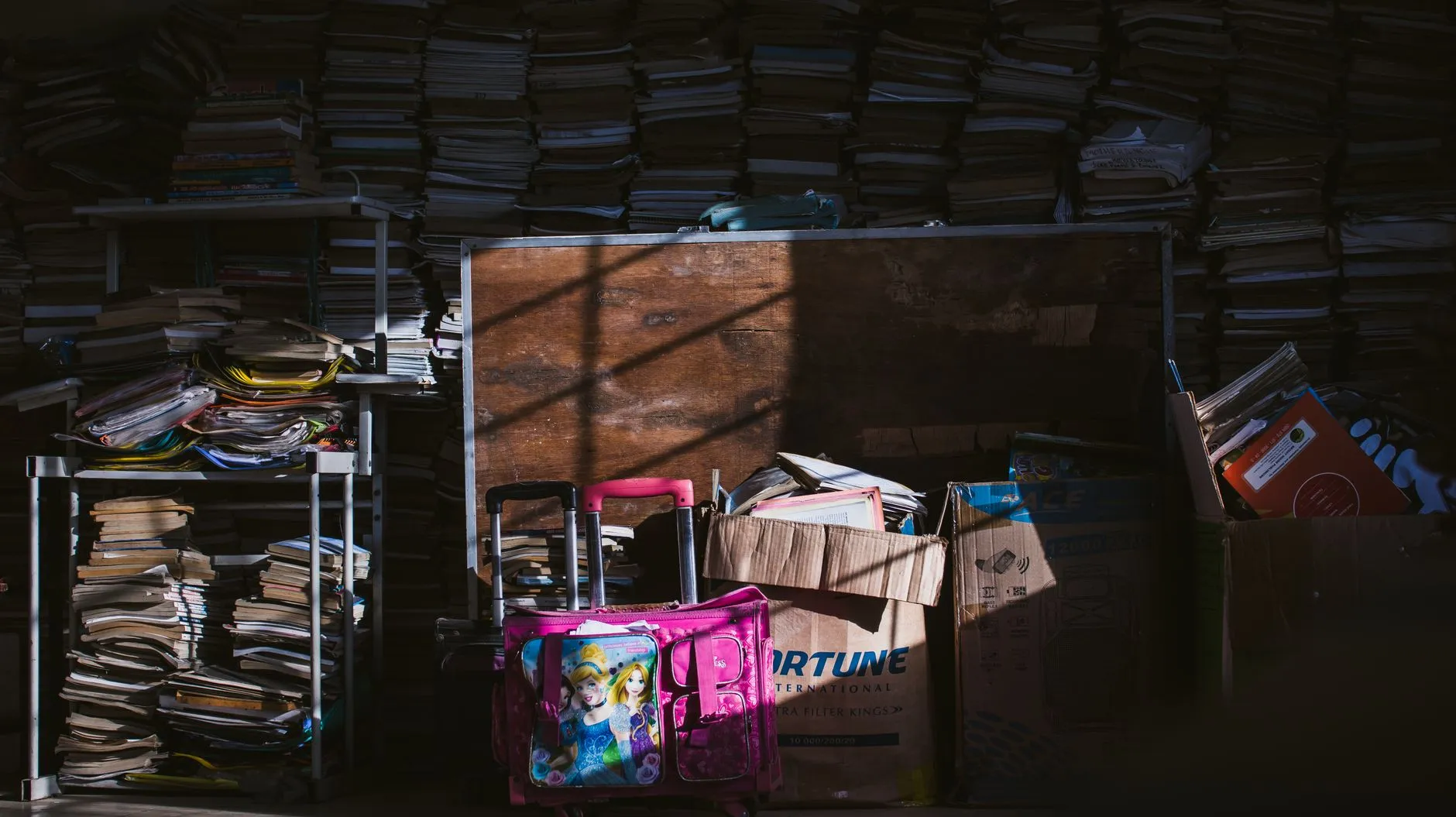 Tumpukan kardus dan buku di ruangan redup dengan bayangan tajam.